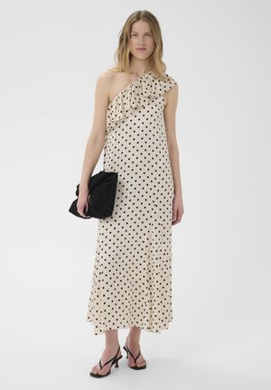 Femme en robe crème une épaule à volants avec des pois noirs, tenant une pochette noire et portant des sandales à talons noires.