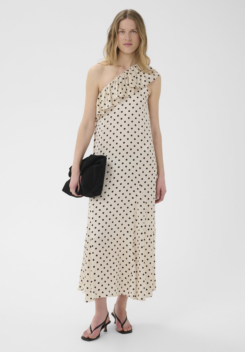 Femme en robe crème une épaule à volants avec des pois noirs, tenant une pochette noire et portant des sandales à talons noires.