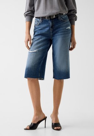 Femme portant un short en jean déchiré mi-long, un haut gris à manches longues, une ceinture noire et des mules noires à talons hauts et bouts ouverts, debout.