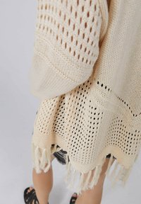 Gesponnener Lochstrick-Poncho in Cremefarbe mit Fransen am Saum und lockerem, entspanntem Schnitt. Durchgehend mit strukturiertem Muster.