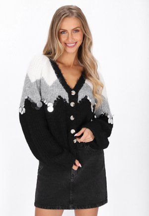 Femme souriante aux longs cheveux blonds portant un cardigan noir, gris et blanc boutonné avec des paillettes et une jupe en denim noire sur fond blanc.