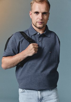 Mężczyzna z krótkimi blond włosami i brodą, ubrany w niebieską koszulkę polo i jasne dżinsy, trzymający czarny pasek plecaka na ramieniu.