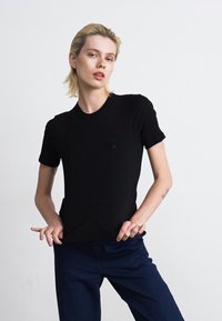 Person med kort, blond hår iført en sort t-shirt og marineblå bukser, stående med hænderne nær hoften mod en ensfarvet hvid baggrund.