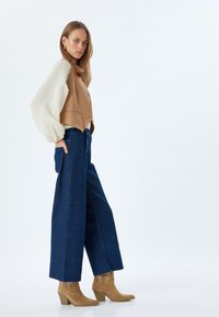 Harmaa hihaton liivi napilla valkoisen, ilmavan pitkän hihansuun paidan päällä, yhdistettynä leveisiin sinisiin farkkuihin ja vaaleisiin korkokenkiin.