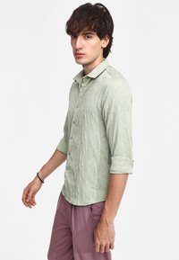 Camisa abotonada verde claro con un patrón geométrico texturizado, mangas largas enrolladas hasta los codos y un diseño de cuello clásico.