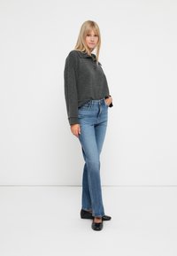 Mørkegrå kortermet genser med krage-design og knapp, parret med lyseblå utsvingte jeans og svarte flate sko. Enkel bakgrunn.