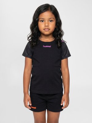Ung pige med sort krøllet hår iført en sort sportst-shirt med lilla logo og sorte shorts med orange logo, stående mod en ensfarvet baggrund.