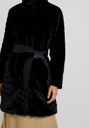 Femme portant un manteau en fausse fourrure noire avec une large ceinture noire nouée à la taille, par-dessus une jupe beige.