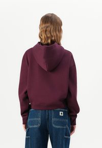 Felpa a maniche lunghe in tessuto bordeaux scuro, con cappuccio regolabile e vestibilità morbida. Indossata con jeans in denim blu, con dettagli sulle tasche posteriori.