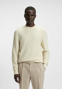 Maglione a maglia color crema con un motivo a waffle testurizzato, colletto rotondo e polsini a coste, abbinato a pantaloni plissettati beige chiaro.