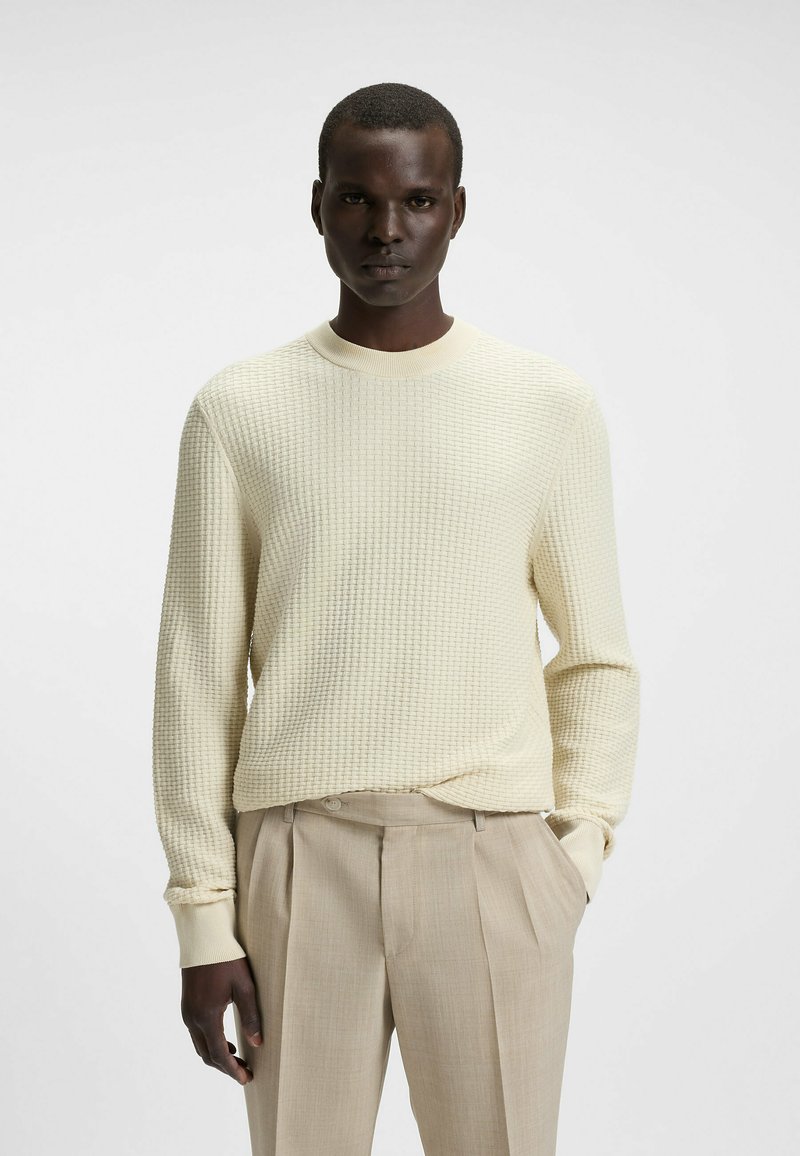 Maglione a maglia color crema con un motivo a waffle testurizzato, colletto rotondo e polsini a coste, abbinato a pantaloni plissettati beige chiaro.