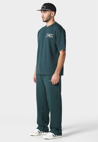 Grønn bomull T-skjorte med brodert tekst og matchende brede joggebukser. Hvite sneakers med marineblå og grønne striper. Svart caps.