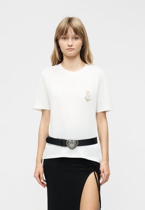 Ung kvinde står med fronten mod kameraet iført hvid t-shirt med lille blomstermotiv, sort bælte med hjerteformet spænde og sort nederdel med sideslids.