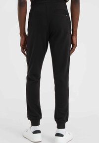 Schwarze Jogginghose mit eng anliegenden Bündchen, aus weichem Baumwollstoff gefertigt. Enthält eine Gesäßtasche; kombiniert mit weißen Sneakers und strukturierten Socken.