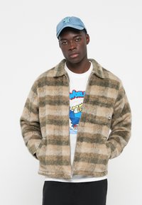 Plaid zip-up jacket in beige and green hues, made from textured fabric. Worn over a graphic white t-shirt with a crew neckline.