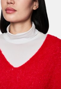 Roter, samtiger Pullover mit V-Ausschnitt, der über einem weißen Rollkragenpullover getragen wird. Der Pullover hat eine strukturierte Oberfläche und ein weiches Erscheinungsbild.