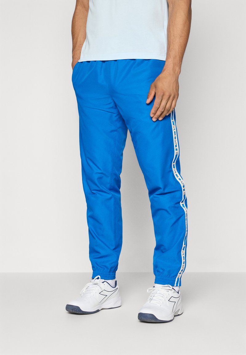 Blauwe sportieve broek gemaakt van lichtgewicht materiaal met een tapse ontwerp. Voorzien van witte zijstrepen met logo-details en elastische boorden.