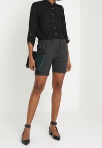Chemise noire boutonnée, shorts gris sur mesure, et escarpins noirs à bride chevillère. Une pochette verte texturée apporte un contraste. Tissu lisse et design soigné.