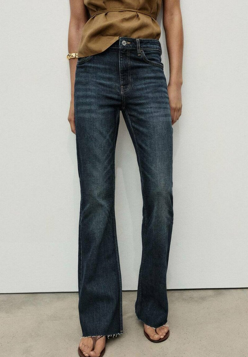 Vaqueros acampanados de color azul oscuro con cintura alta. El material es denim con un acabado ligeramente desgastado. Presenta un dobladillo sin terminar y bolsillos frontales.
