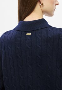 Μπλε ναυτικό πουλόβερ με πλέξη καλωδίου και υφή, με ριμπ γιακά και μια μικρή χρυσή πλακέτα με λογότυπο στο πίσω μέρος.