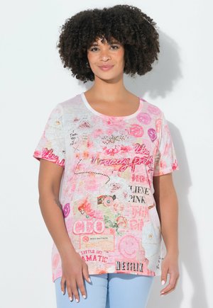 Kobieta z kręconymi włosami, ubrana w kolorowy, wzorzysty różowo-biały T-shirt i jasnoniebieskie spodnie, stojąca na białym tle.