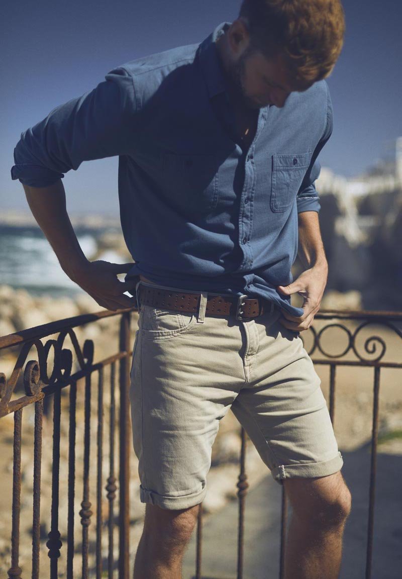Lichtblauw overhemd met lange mouwen, opgerolde mouwen, gecombineerd met crèmekleurige shorts en een bruine riem, staand bij een reling met uitzicht op de zee.