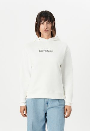 Vit huvtröja med huva som har "Calvin Klein"-logotypen i svart, tillverkad av mjukt tyg med en avslappnad passform, kombinerad med ljusblå denimbyxor.