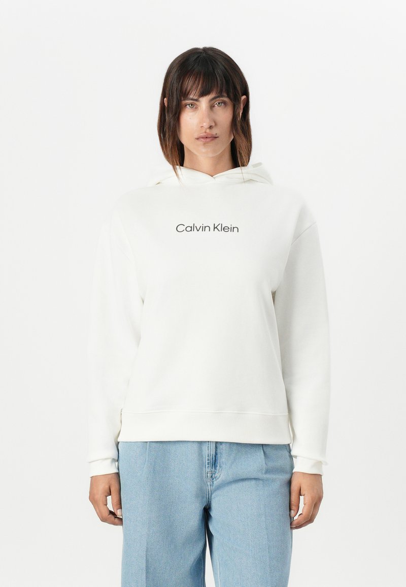 Felpa bianca con cappuccio che presenta il logo "Calvin Klein" in nero, realizzata in tessuto morbido e con una vestibilità comoda, abbinata a pantaloni in denim blu chiaro.