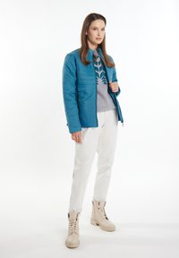 Jachetă teal, matlasată, cu guler înalt, asortată cu un pulover gri deschis cu un model de frunze verzi și pantaloni albi. Bocanci cremi.