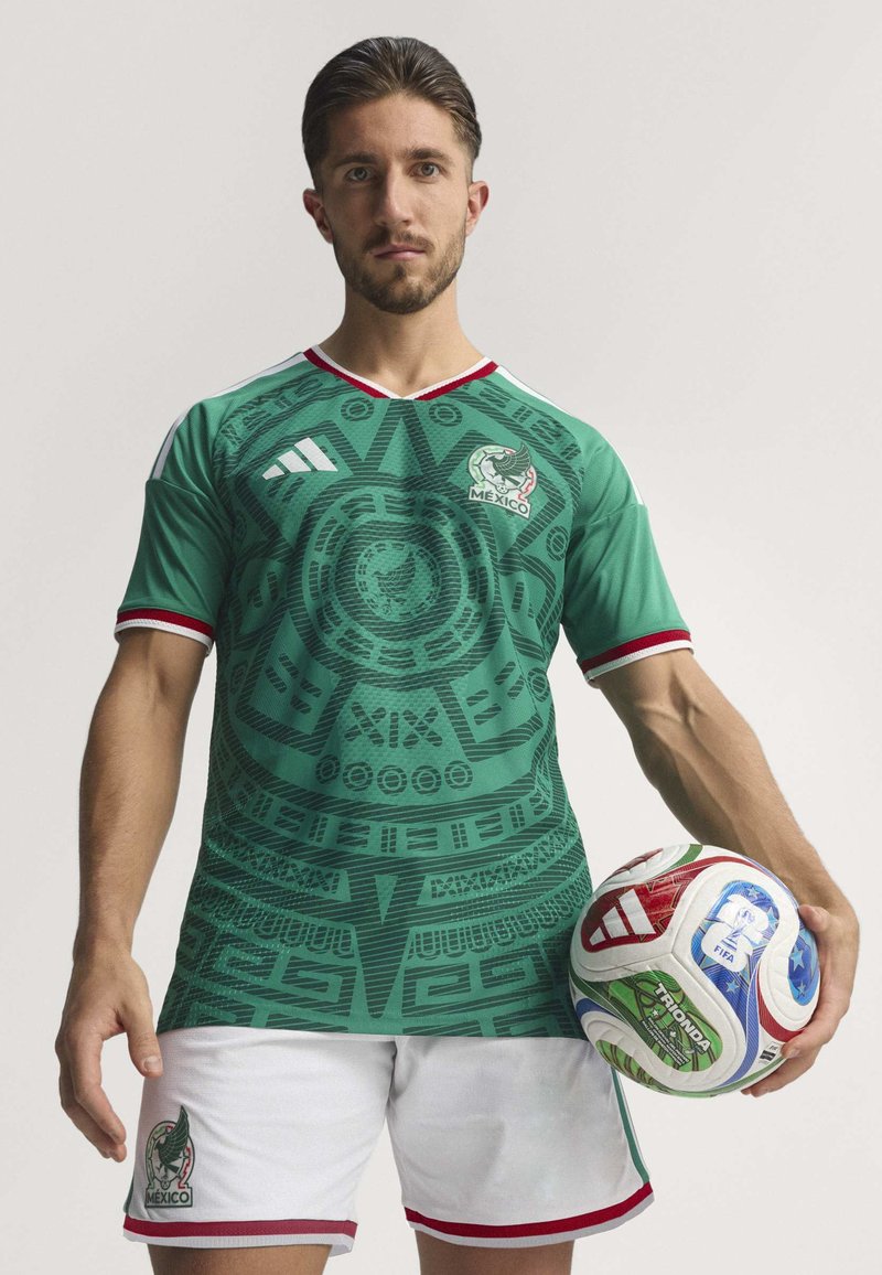 Green soccer jersey with textured Aztec-inspired design, red accents on the collar and sleeves, paired with white shorts and a multicolored soccer ball.