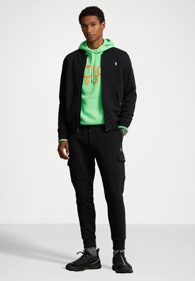 Sort zip-up bomberjakke over en lys grøn hoodie med orange grafik, parret med sorte joggingbukser og sorte sneakers med strukturerede detaljer.