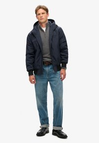 Marinblå jakke med hætte over en grå sweater. Blå jeans med opsmøg og sorte sko. Hverdagsoutfit med funktionelt design og lynlåsdetaljer.
