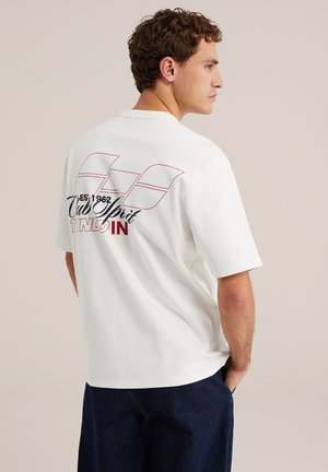 Man met krullend haar draagt een wit oversized T-shirt met meerkleurige grafische tekst op de rug, gecombineerd met donkerblauwe broek, handen in de zakken.