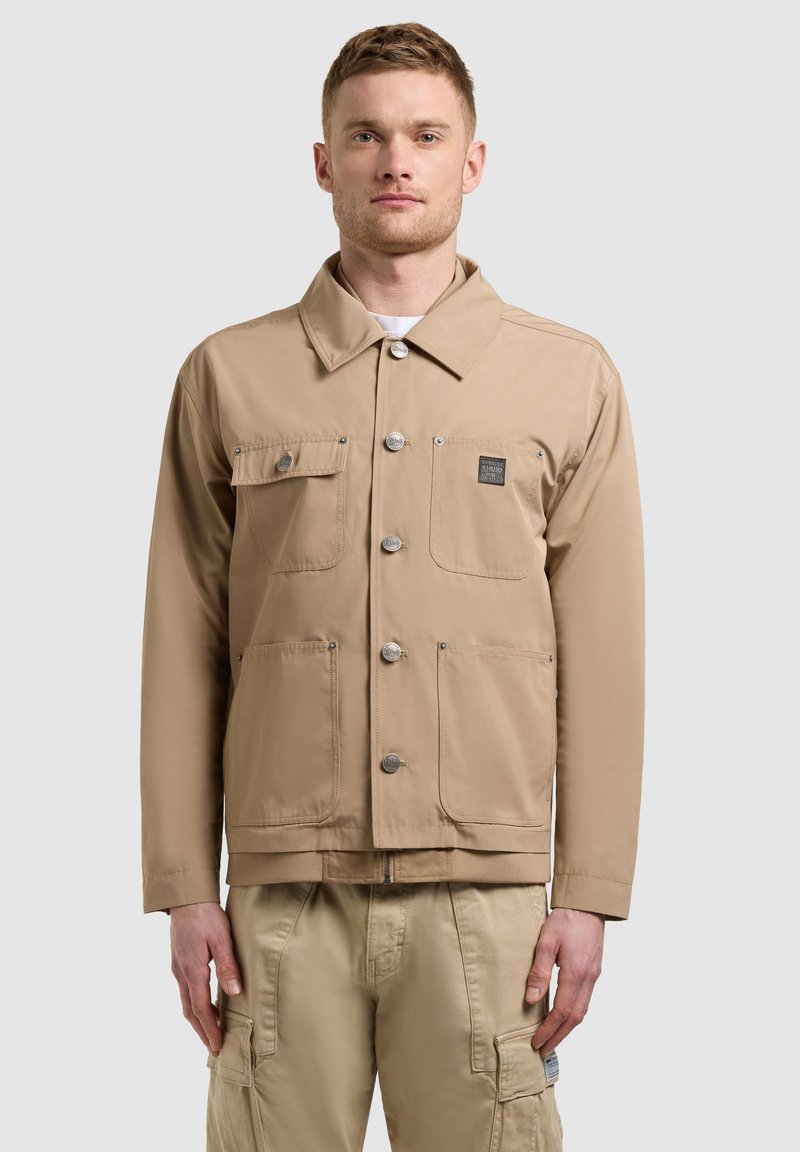 Homme portant une veste beige boutonnée avec quatre poches avant et un pantalon cargo beige assorti, debout devant un fond uni.