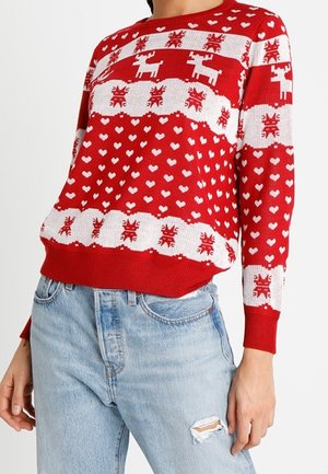 Pull en tricot rouge avec des motifs de rennes et de cœurs blancs, des poignets et un ourlet côtelés, et une coupe décontractée. Porté avec un jean en denim usé.
