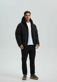 Schwarze gepolsterte Jacke mit Kapuze, über einem weißen T-Shirt getragen, kombiniert mit schwarzen Cargo-Hosen und braunen Turnschuhen mit blauen Schnürsenkeln.