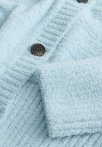 Tissu bleu clair texturé avec un motif côtelé, comportant un bouton marron foncé à quatre trous. Gros plan montrant les détails du matériau et des coutures.