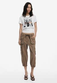T-shirt blanc avec un graphique de chat et du texte, associé à un pantalon cargo marron avec de grandes poches et des poignets élastiques, porté avec des sandales noires à lanières.
