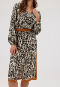 Femme portant une robe noire et beige à motif abstrait avec des manches bouffantes, une taille marquée et une bordure marron à l’ourlet et sur les côtés.