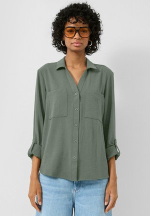 Kobieta z kręconymi włosami, nosząca duże bursztynowe okulary przeciwsłoneczne, srebrne kolczyki-koła, oliwkowo-zieloną koszulę zapinaną na guziki oraz jasnoniebieskie jeansy na jednolitym tle.