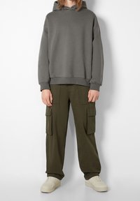 Persona che indossa un felpa grigia con cappuccio, pantaloni cargo verde oliva con tasche laterali e sneakers color crema, in piedi contro uno sfondo bianco semplice.
