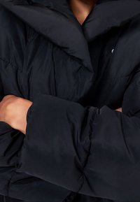 Sort pufferjakke med høj krave, oversized design, quiltet tekstur og brede ærmer. Har en glat, shiny stoffinish.