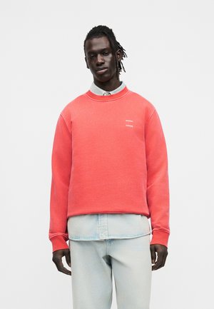 Homme portant un sweat corail sur une chemise en denim clair et un jean, debout devant un fond blanc uni.