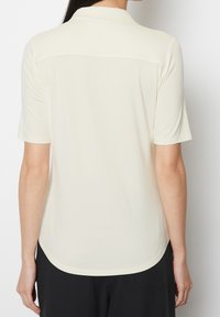Jasno beżowa koszulka polo z krótkim rękawem, gładka w dotyku, z tylną karczkiem i zaokrąglonym dołem. Prosty design, odpowiednia do codziennego noszenia.