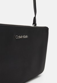 Juodas odinis rankinis krepšys su tekstūruota paviršiumi, su sidabrinės spalvos Calvin Klein logotipu ir užtrauktuku viršuje.