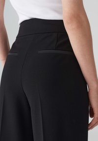 Pantalon noir taille haute en tissu lisse, doté de deux petites poches arrière au design minimaliste et sans quincaillerie visible.