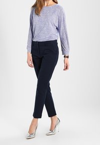 Ljusblå långärmad topp med texturerad tyg, tillsammans med mörkblå tajta byxor och silvriga högklackade skor, stående mot en neutral bakgrund.