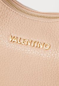 Bež torba iz drobnozrnate usnje z zlatim okovjem. Ima vidno zadrgo in vtisnjen logo "VALENTINO" na sprednji strani.