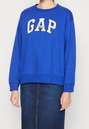 Osoba nosząca jasno niebieską bluzę GAP z długim rękawem, w połączeniu z ciemną spódnicą dżinsową, na jednolitym jasnym tle.