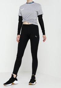 Camiseta gris anudada con mangas largas negras, mallas negras con un pequeño logo de Puma y zapatillas negras con acentos dorados.
